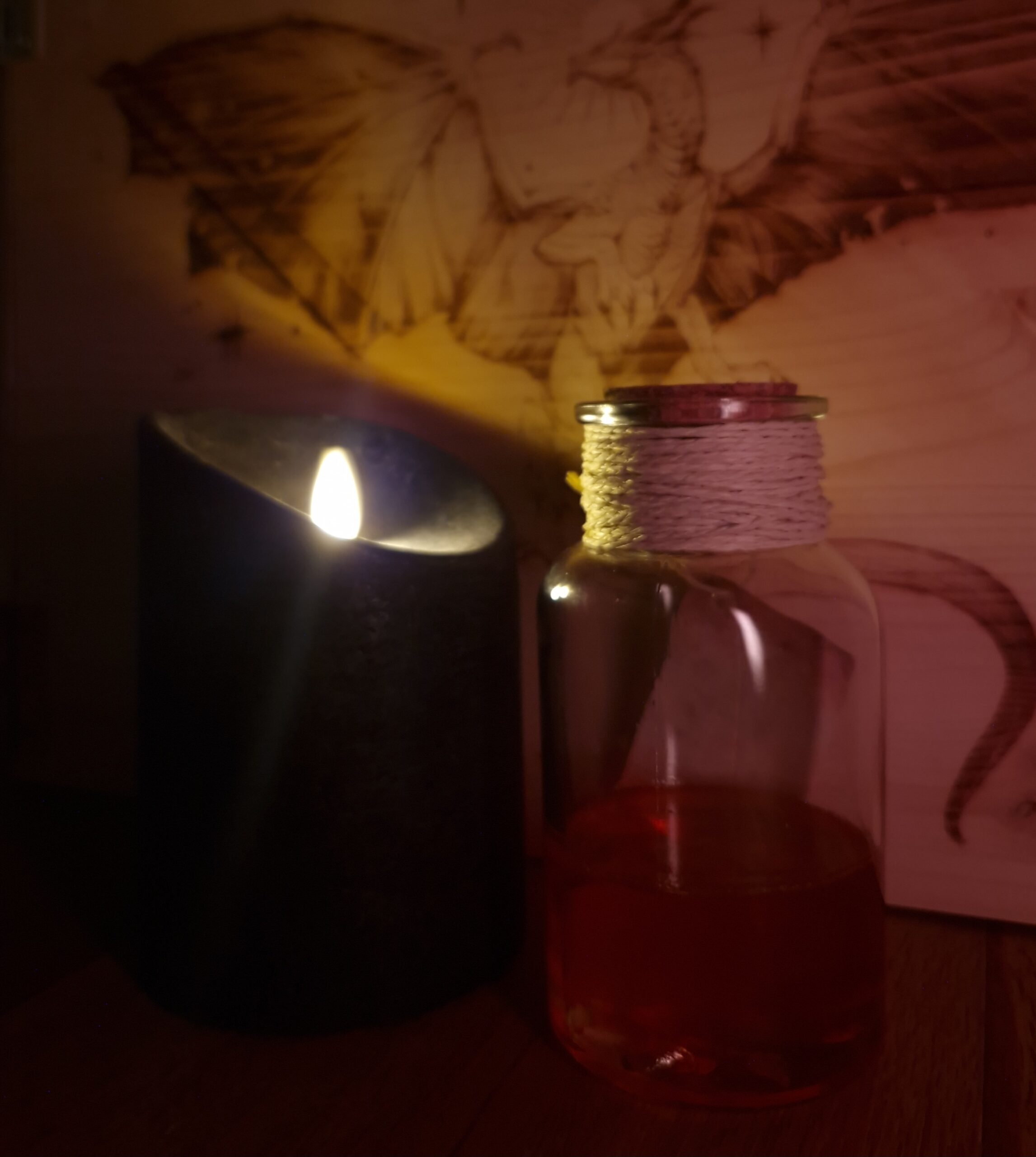 Eine Glasflasche mit Korken und bast um den halb mit einer roten Flüssigkeit darin. Daneben steht eine Schwarzen Kerze. Im Hintergrund ist eine Holzwand mit einem eingebrannten Drachen zu sehen.